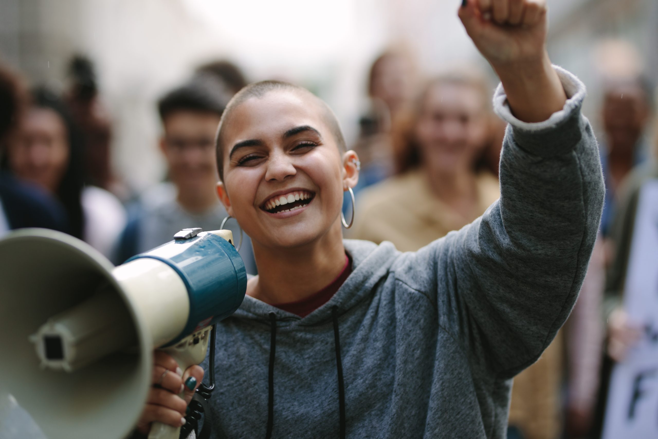A White woman with a shaved head holds up a fist and a megaphone as she smiles