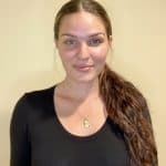 Photo of Irisa MacAulay. She is standing in front of a white wall. She has long brown hair pulled back in a ponytail and is wearing a long sleeve black shirt.
