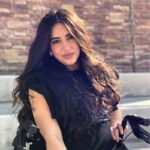 Foto de MaryAngel, mujer con cabello oscuro y largo vistiendo una blusa negra sentada frente a una pared de ladrillos cobrizos.