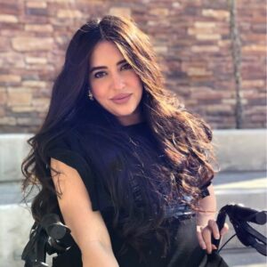 Woman with long, wavy dark hair wearing a black top, sitting outdoors in front of a reddish-brown stone wall.