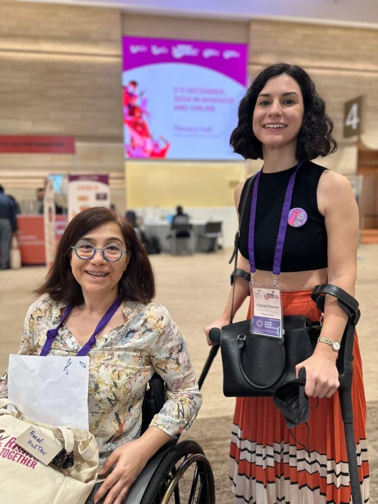 Photograph features Virginia Ossana, a light-skinned woman with dark, curly hair, smiling while wearing a green blouse and a purple lanyard. Virginia uses crutches. Next to her is Amal, an activist from Lebanon who has shoulder lenght brown hair, she is wearing a multi-colored blouse in beige and yellow tones. She is in a wheelchair.