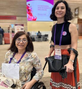 Photograph features Virginia Ossana, a light-skinned woman with dark, curly hair, smiling while wearing a green blouse and a purple lanyard. Virginia uses crutches. Next to her is Amal, an activist from Lebanon who has shoulder lenght brown hair, she is wearing a multi-colored blouse in beige and yellow tones. She is in a wheelchair.