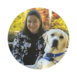Janie poses with her service dog Nevile VII in front of a landscape with trees.