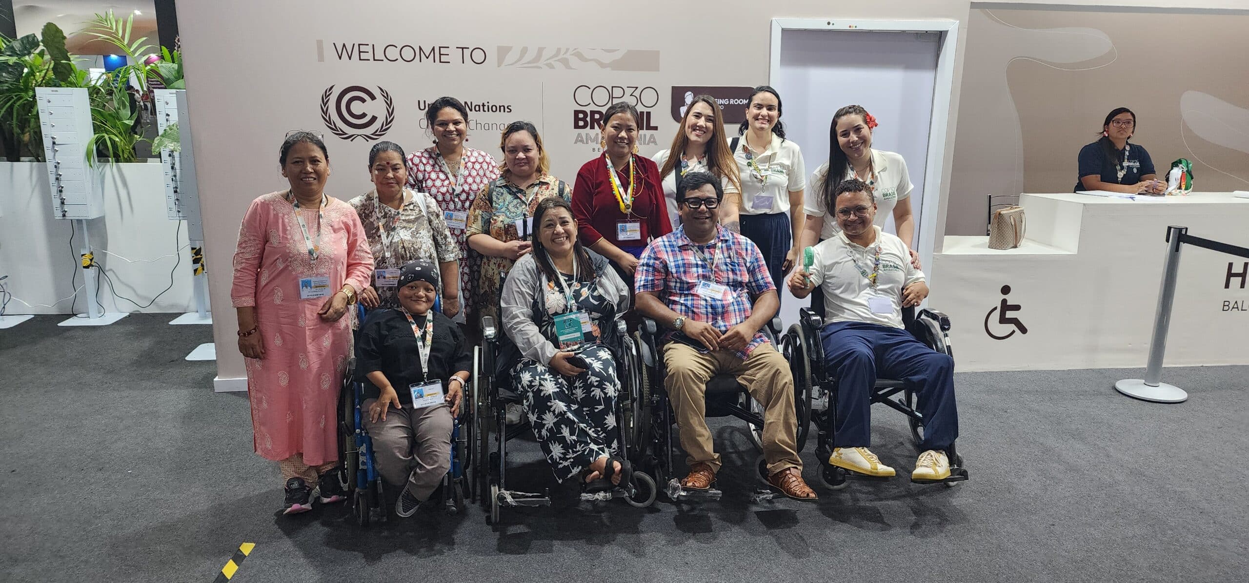 Un grupo de doce personas, entre las que se encuentran varies usuaries de sillas de ruedas, sonríen y posan juntes en la sede de la Conferencia de las Naciones Unidas sobre el Cambio Climático (COP30) en Belém, Brasil. Se encuentran delante de una pared en la que se lee “Welcome to COP30 Brazil Amazonia” (Les damos la bienvenida a la COP30 Brasil Amazonia) con el logotipo de la Convención Marco de las Naciones Unidas sobre el Cambio Climático. El grupo incluye activistas de los derechos indígenas, de género y de las personas con discapacidad que llevan insignias de la conferencia y lucen vestimentas propias de sus culturas. En el fondo, se pueden ver señales de accesibilidad y personal de la conferencia. 