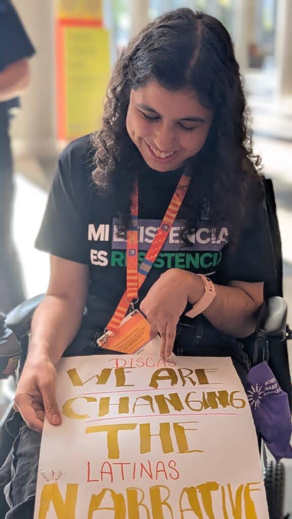 ENG. Gianna, an Argentinian disabled woman and disability advocate is sitting down and holding a sign that says "Disabled woman - we are changing the (latinas) narrative". She is smiling and looking down.