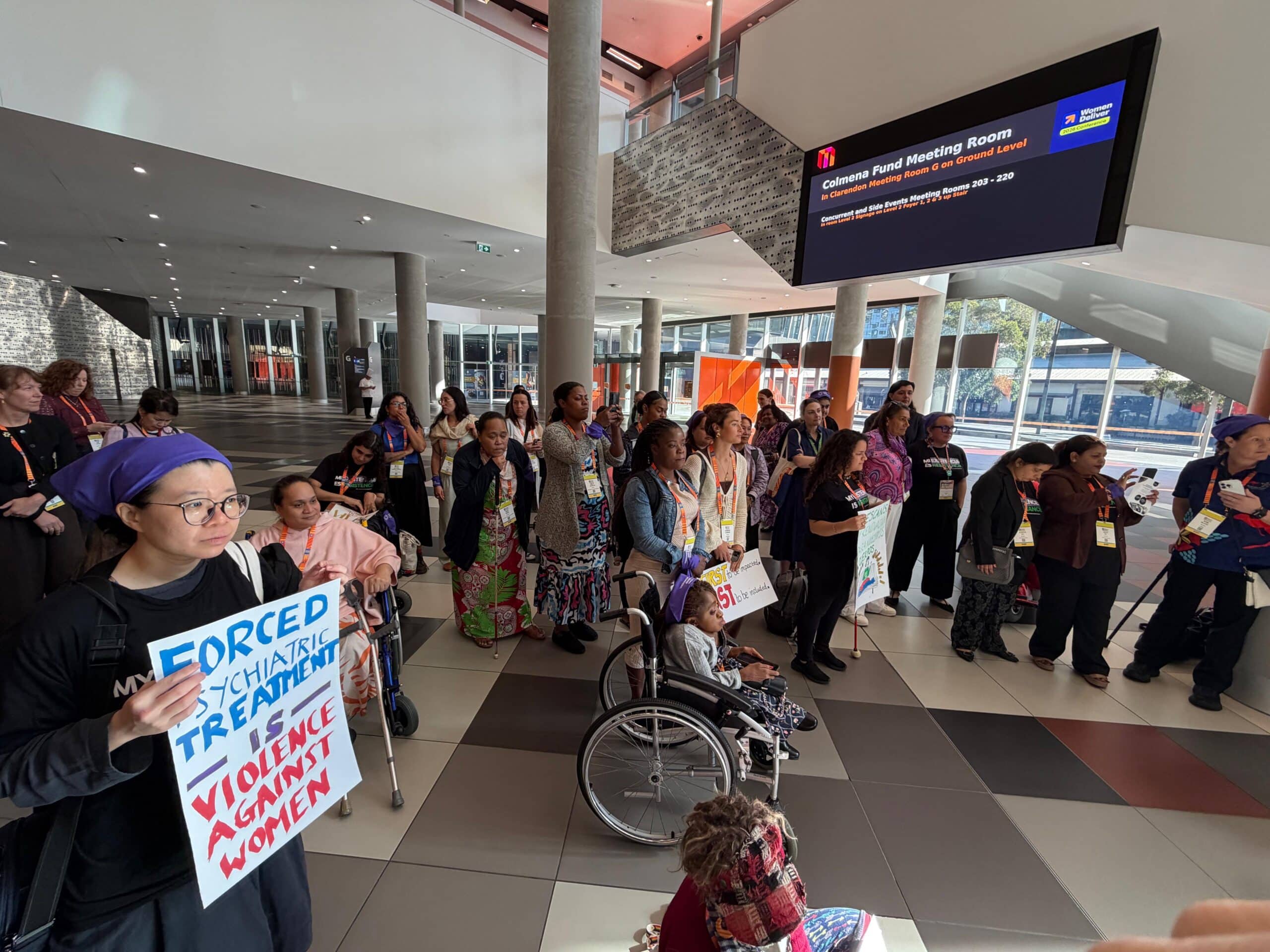 ENG. Women Deliver 2026 women with disabilities meet-up. A diverse group of women with different disabilities are standing and holding signs about Disability Rights and Disability Justice on Women Deliver's 2026 venue.