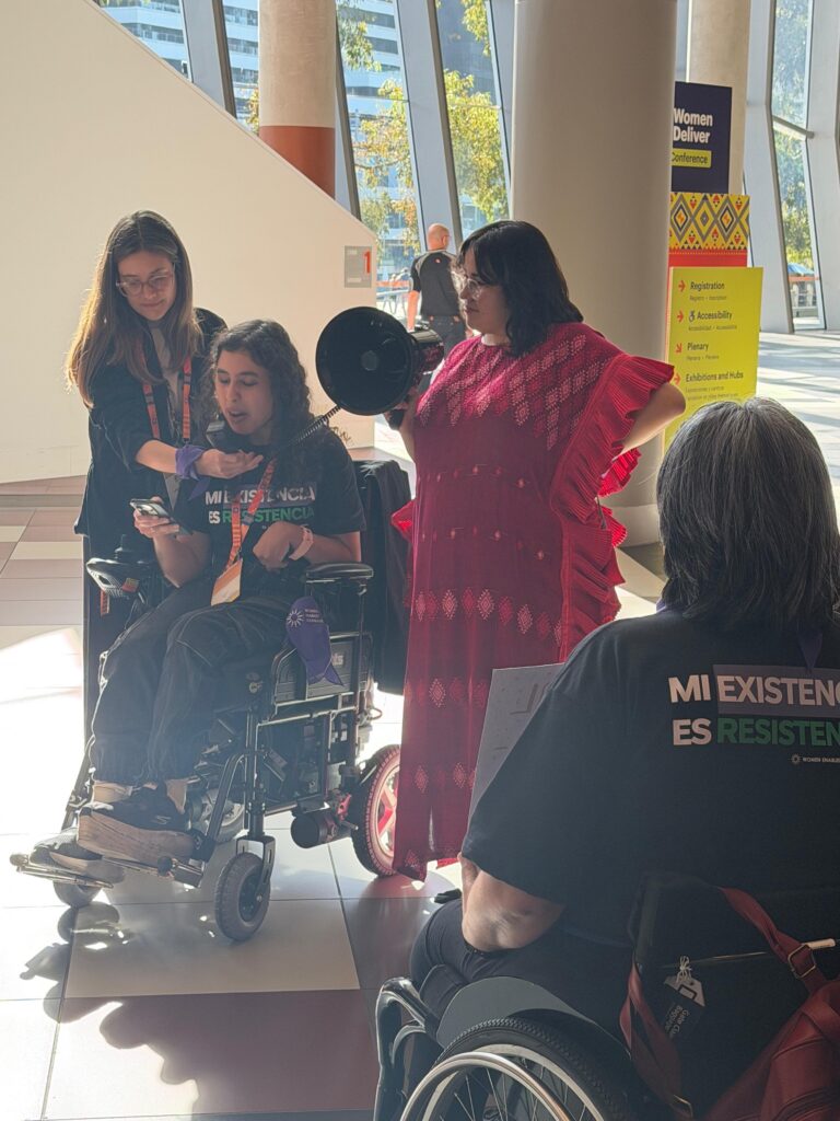 ENG. Gianna Mastrolinardo, an Argentinian disabled woman and disability advocate is speaking through a megaphone and holding her cellphone. She is sitting on her wheelchair. A group of disabled women is near her, some are standing and some are also using wheelchairs. She and others are wearing a t-shirt that reads "My existence is resistance."