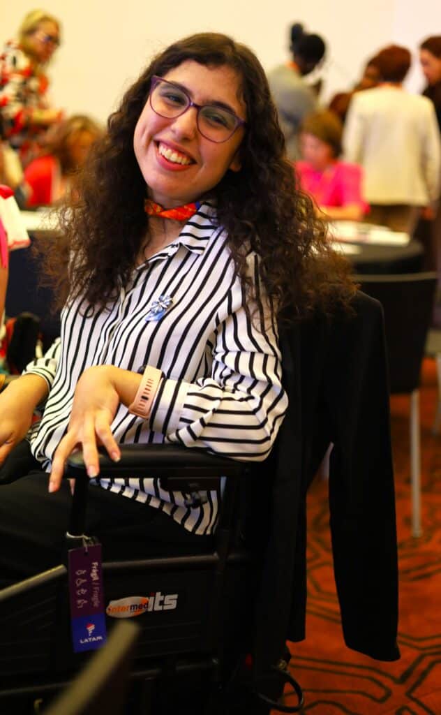 ENG. Image of Gianna Mastrolinardo, a disabled women and disability advocate from Argentina. She is wearing a white shirt with black stripes, has curly long hair and is sitting on an automatic wheelchair.